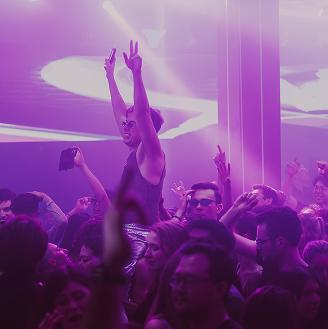 Person raising peace signs in a crowded, purple-lit nightclub.